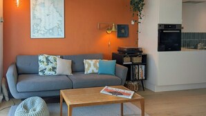 Een woonkamer met een grijze bank, een houten salontafel, een kaart aan de muur, een lamp en een keukenhoek met een waterkoker.