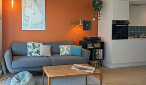 Ein Wohnzimmer mit grauem Sofa, einem Holztisch, einer Karte an der Wand, einer Lampe und einer Küchenzeile mit Wasserkocher.