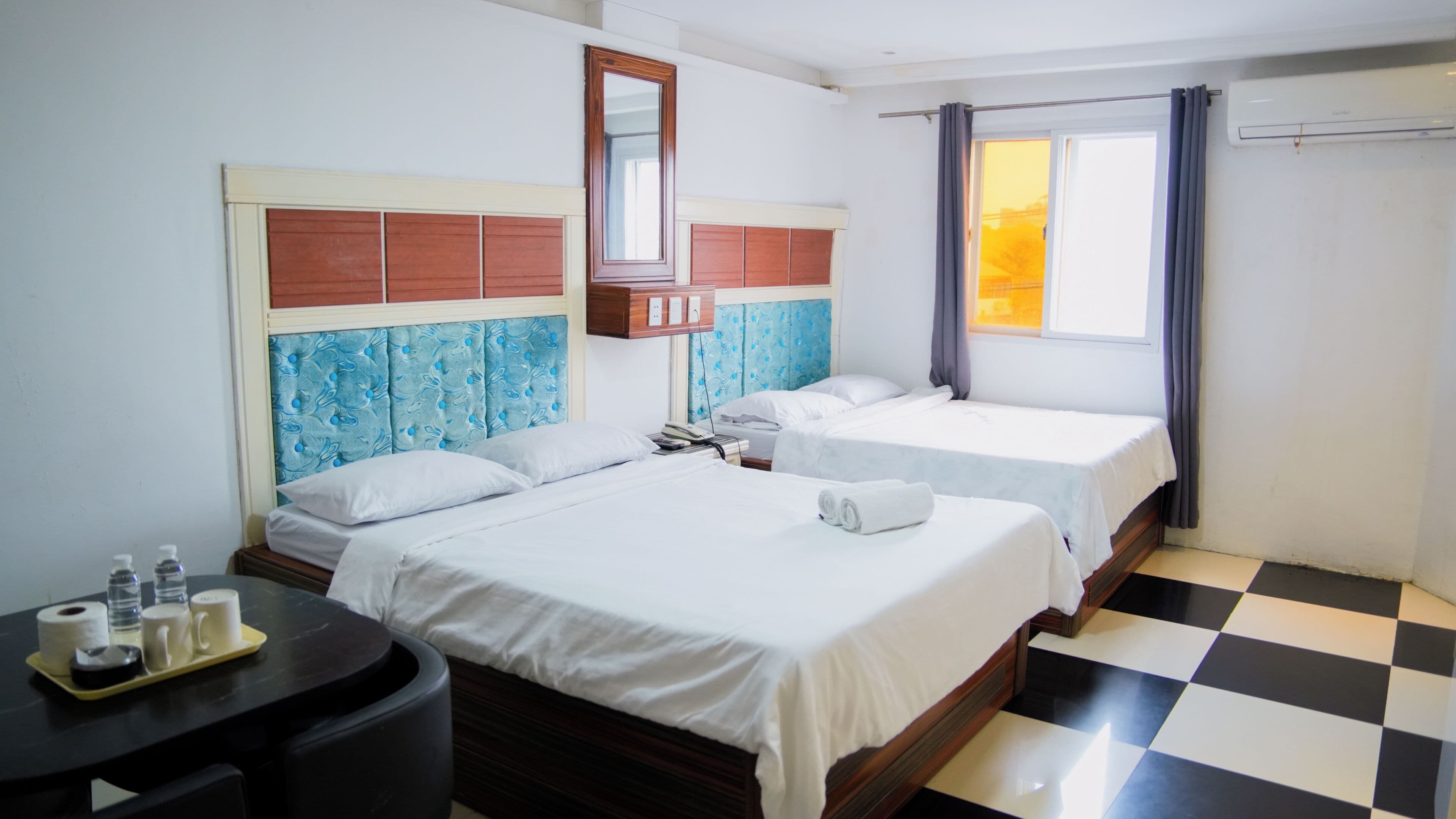 A hotel room with two beds, a desk with toiletries, a mirror, and a window with curtains.