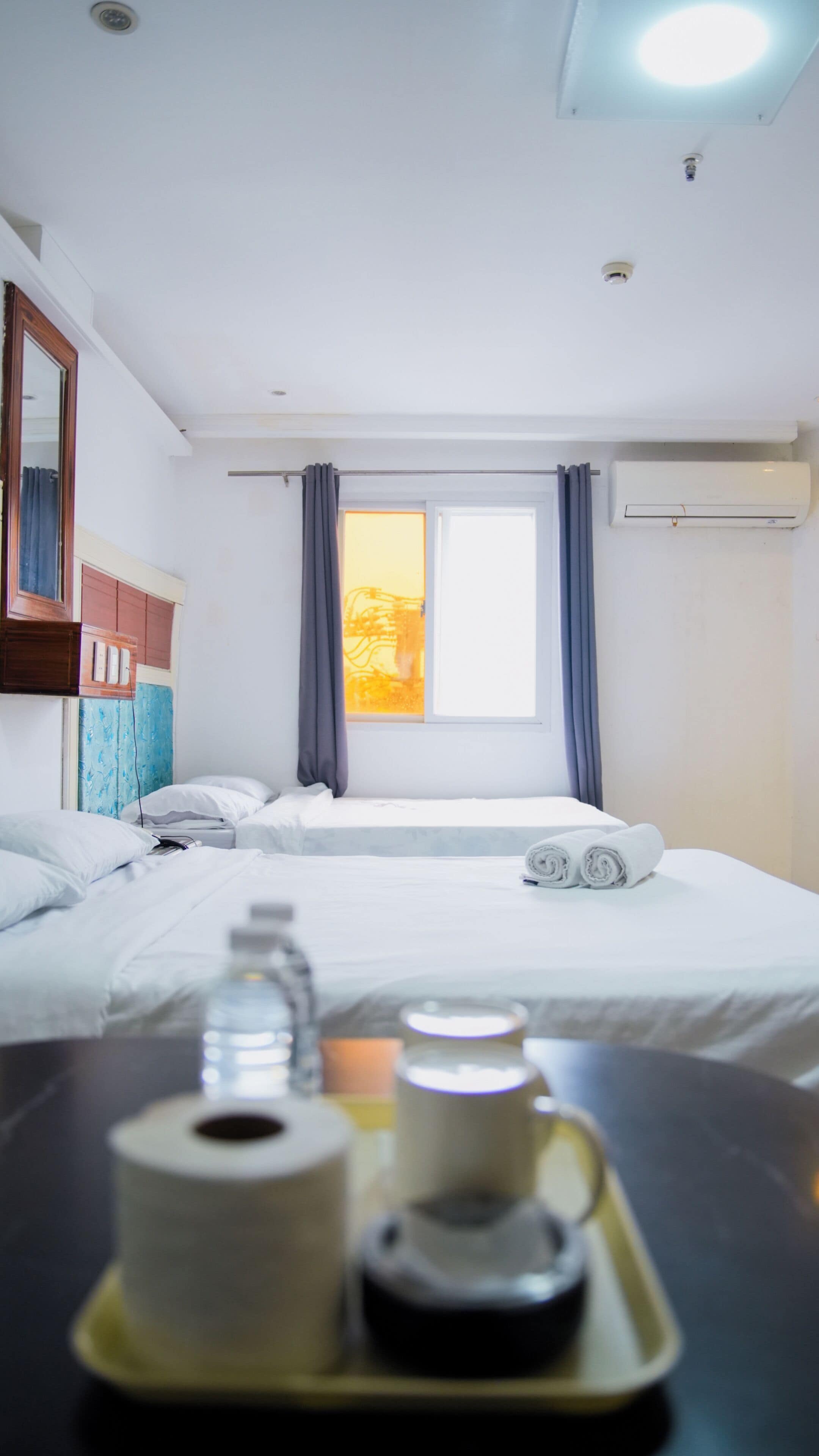 A hotel room with a bed, a window with curtains, an air conditioning unit, and a tray with a bottle and a mug.
