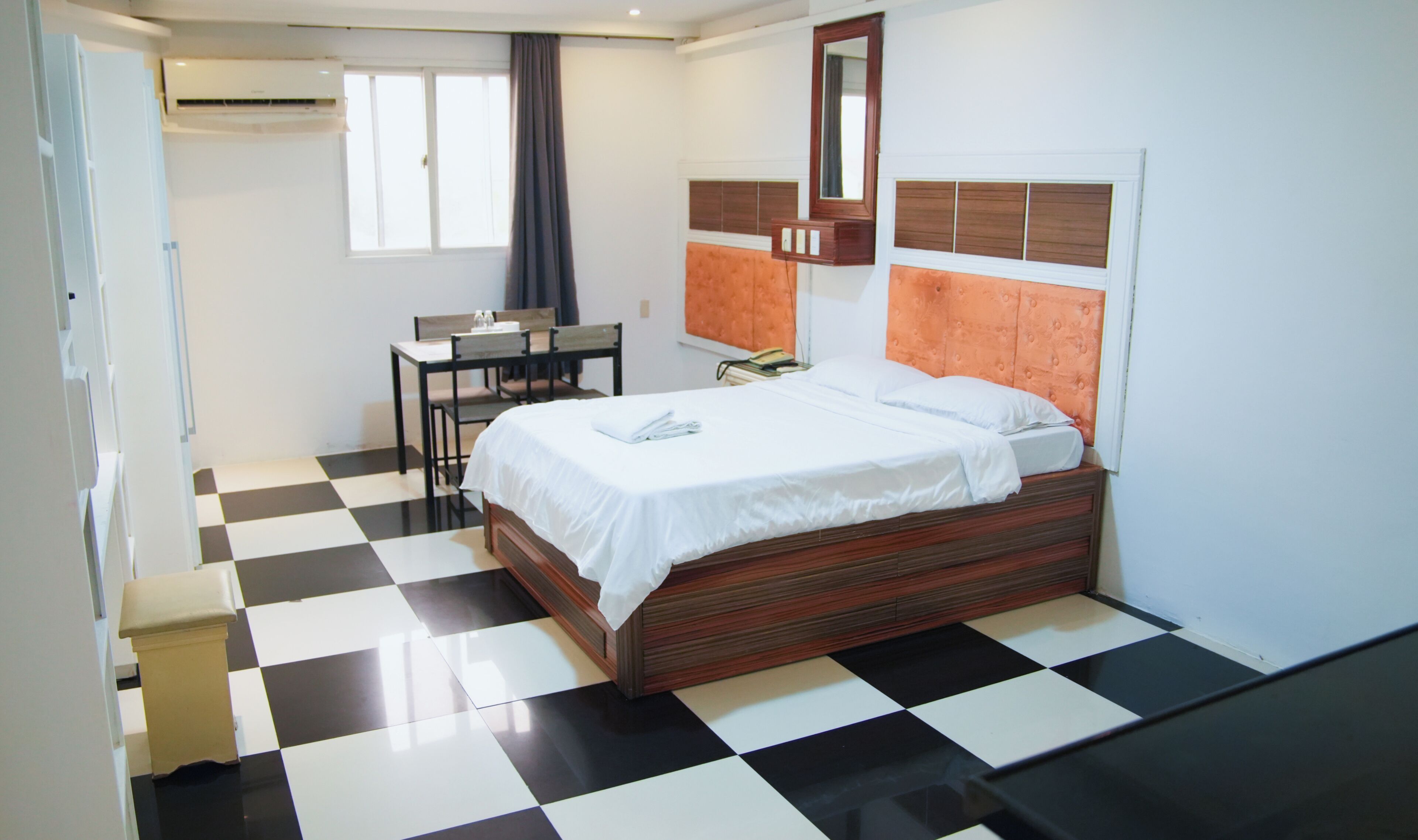 A hotel room with a checkered floor, a bed with a wooden headboard, a desk with a chair, and a window with curtains.