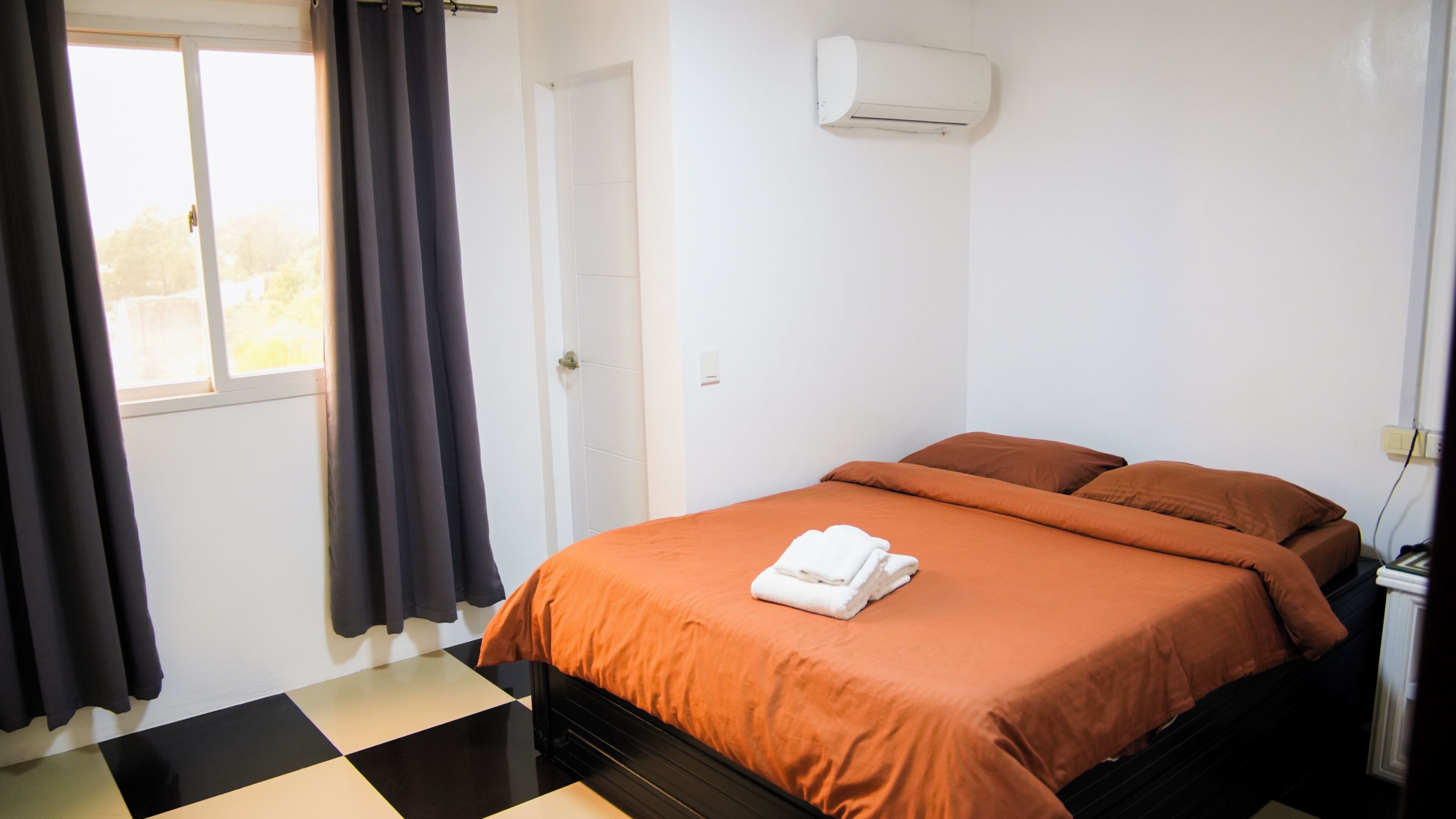 A room with a bed, a window with curtains, a wall-mounted air conditioner, and a checkered floor.