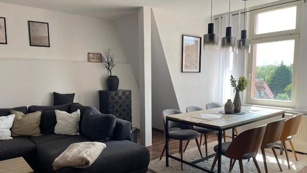 Ein modernes Wohnzimmer mit einem dunklen Sofa, einem Esstisch mit Stühlen, einem großen Fenster und Blick ins Grüne.