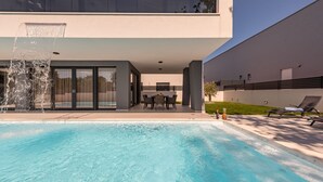 Una piscina moderna con fuente de agua, mobiliario exterior y diseño arquitectónico minimalista.