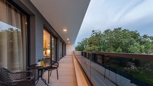 Un balcón con barandilla de vidrio, una mesa con sillas y vista a árboles.