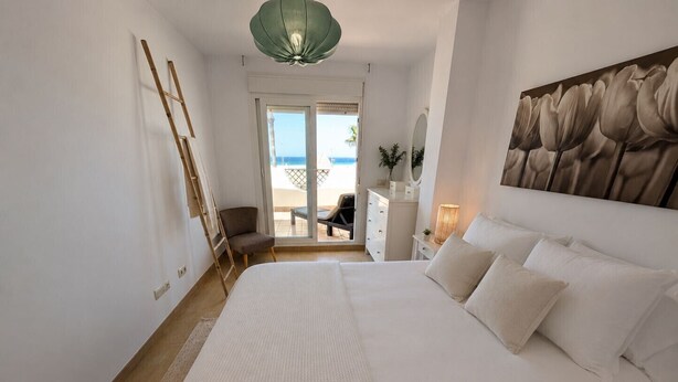 Una camera da letto con un letto, una sedia, una cassettiera, una lampada e vista sulla spiaggia.