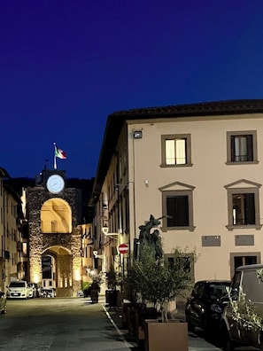 Una strada al crepuscolo con una torre dell'orologio, auto parcheggiate e edifici con finestre ad arco.
