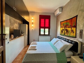 A bedroom with a bed, bedside table, wall-mounted air conditioner, window with red blind, and a painting on the wall.