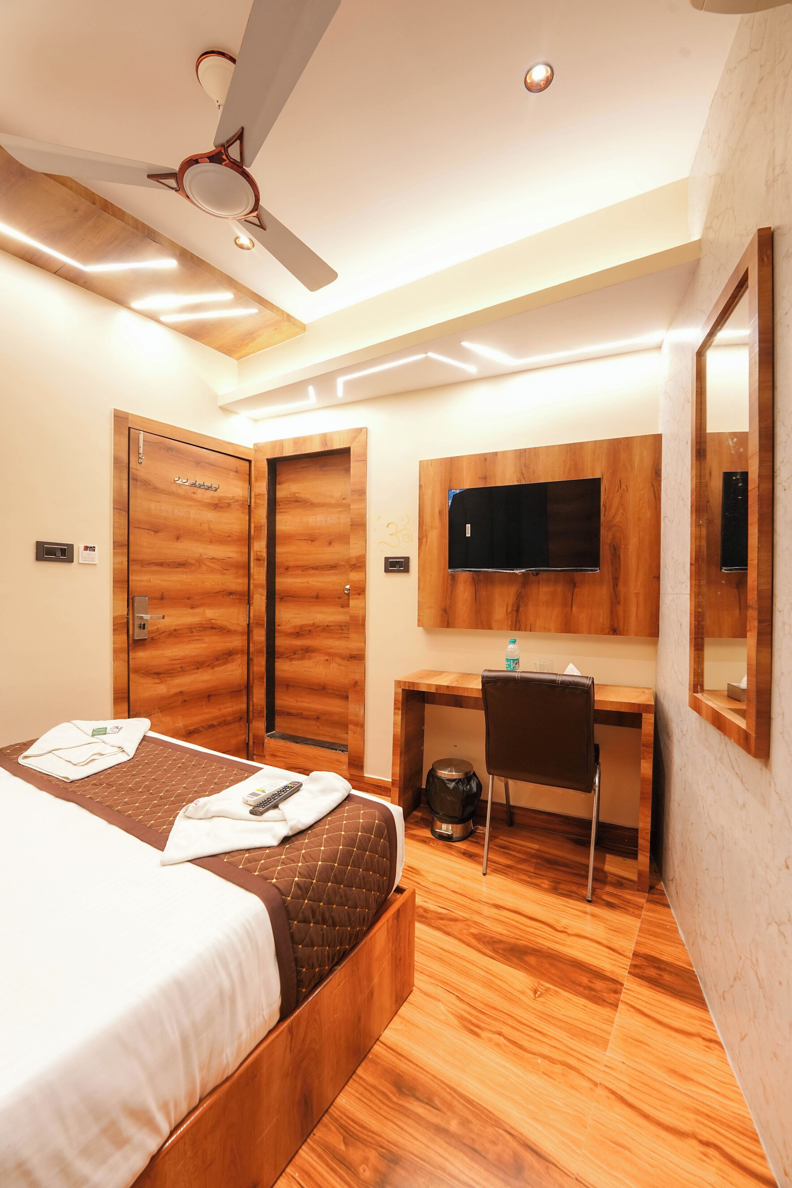 A hotel room with a bed, desk, chair, ceiling fan, TV, and wooden flooring.