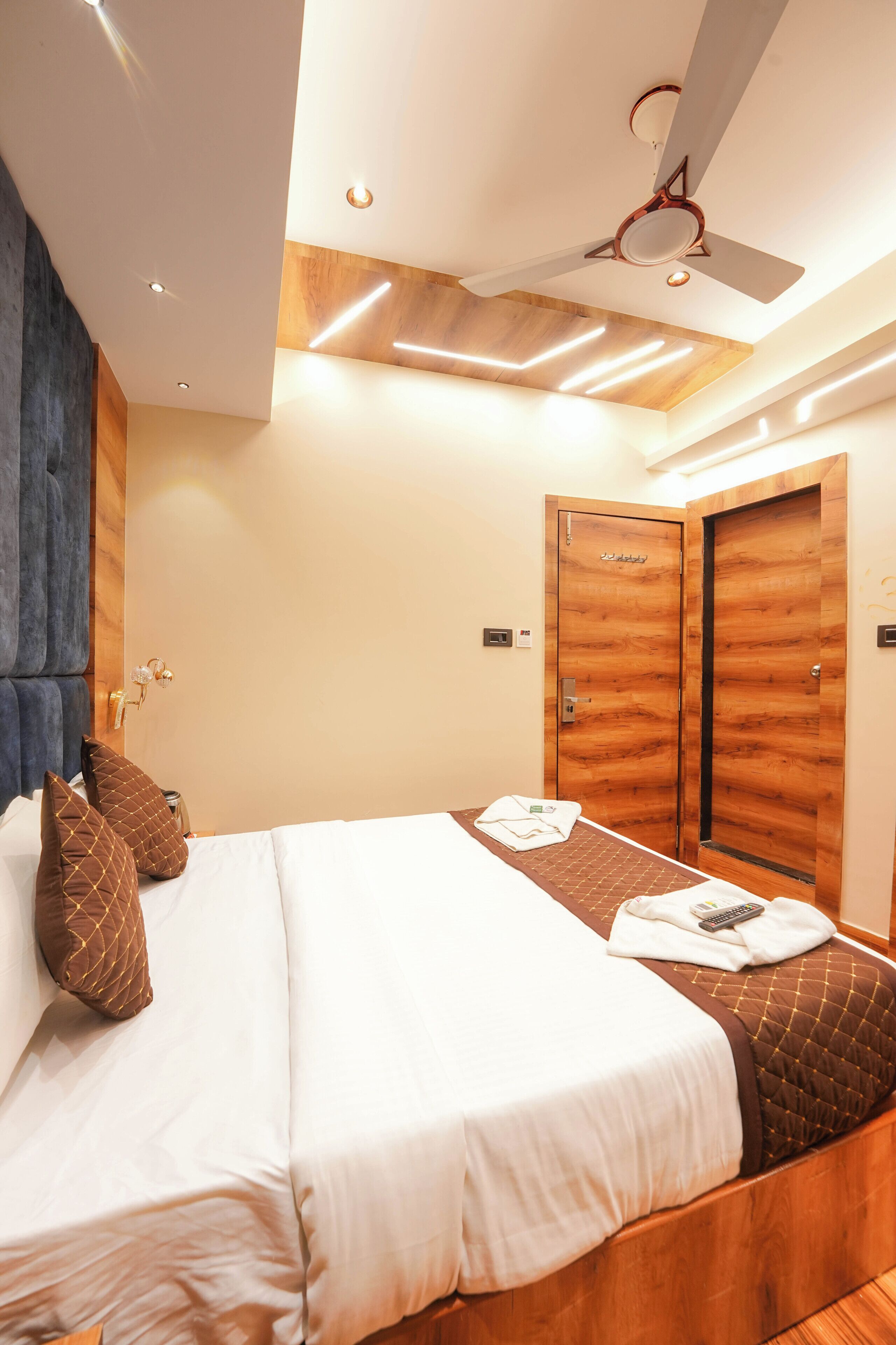 A modern hotel room with a large bed, wooden headboard, ceiling fan, recessed lighting, and wooden doors.