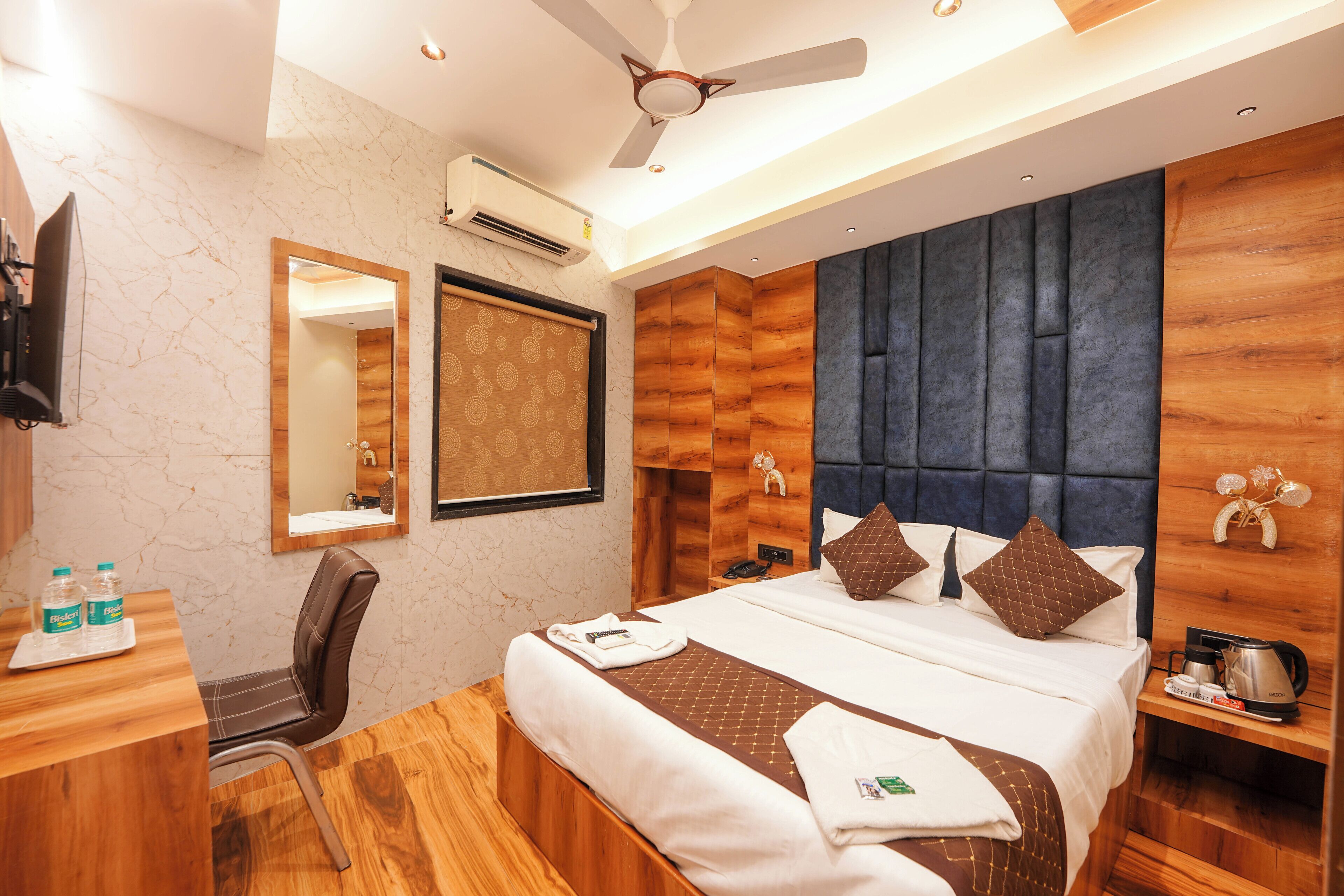 A hotel room with a bed, a desk with a chair, a mirror, a ceiling fan, and a wall-mounted air conditioner.