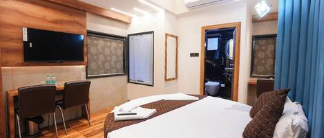 Ein Hotelzimmer mit Bett, Schreibtisch mit Stühlen, einem an der Wand befestigten Flachbildfernseher und einem Fenster mit Vorhang.