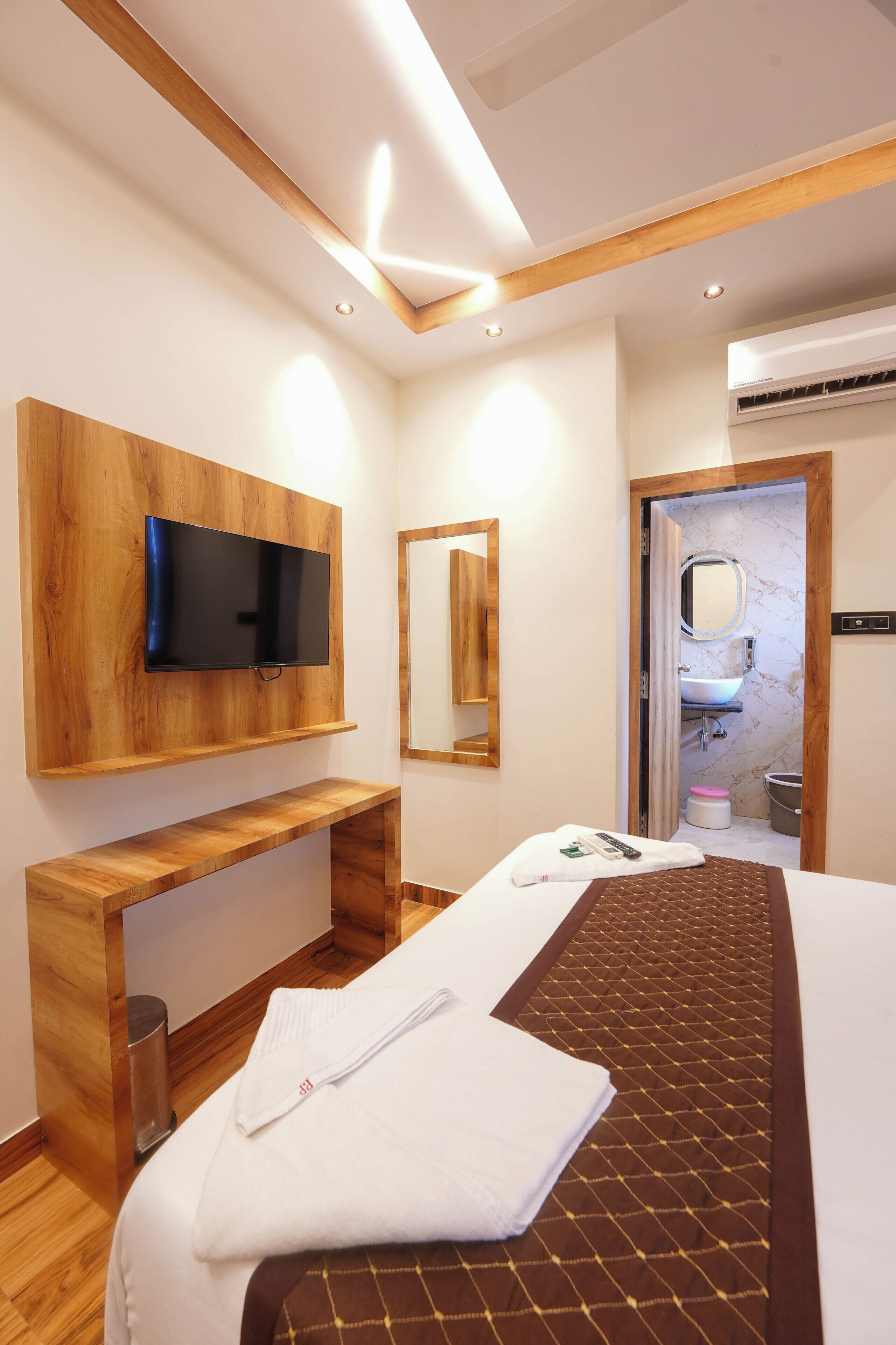 Habitación de hotel con cama, televisor montado en un panel de madera, espejo y baño visible a través de una puerta abierta.