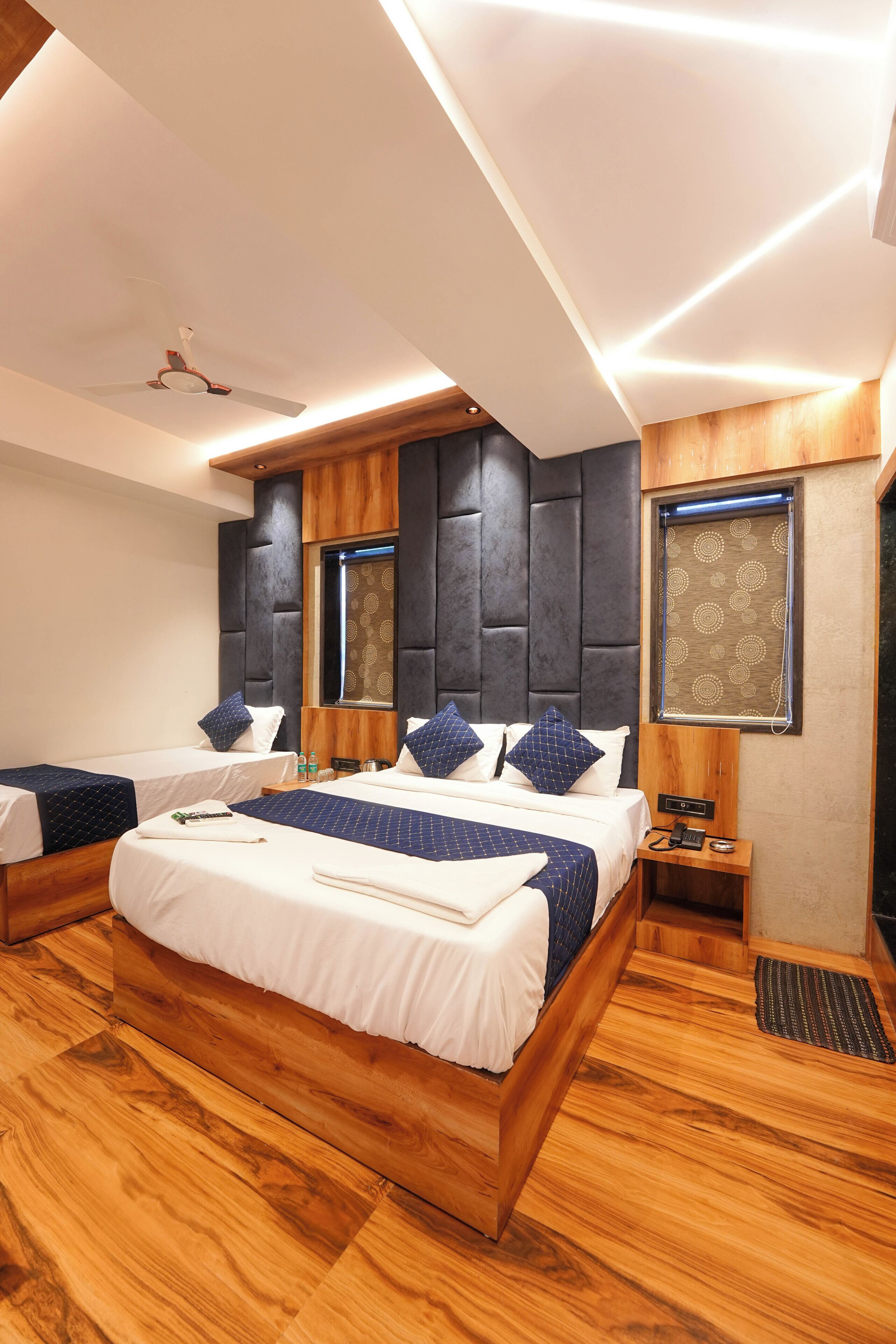 A modern hotel room with a large bed, two smaller beds, wooden flooring, a ceiling fan, and a window with a patterned blind.