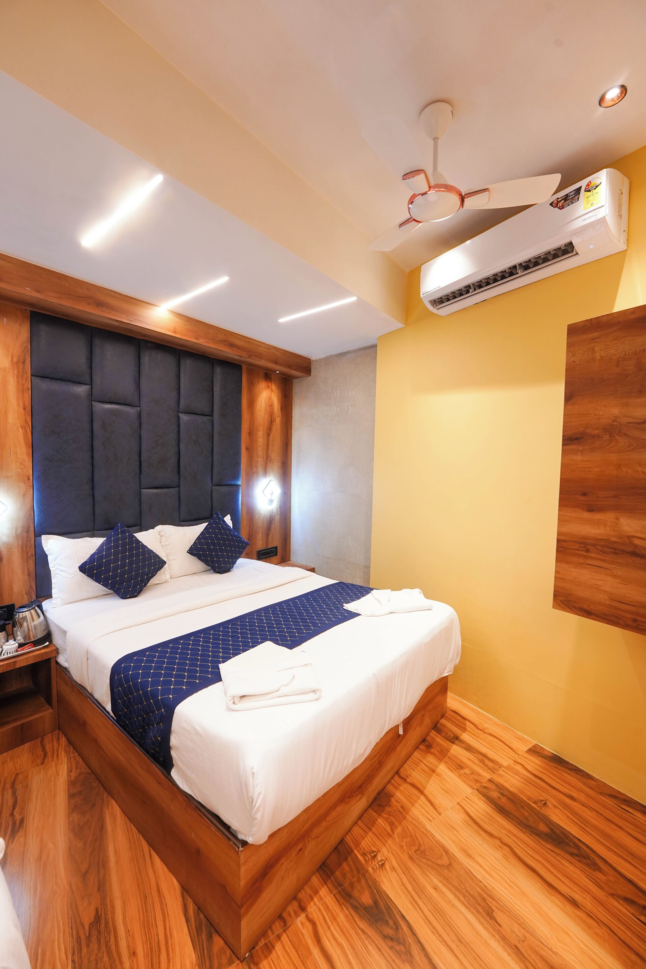 A hotel room with a bed, wooden headboard, ceiling fan, air conditioning unit, and wooden flooring.
