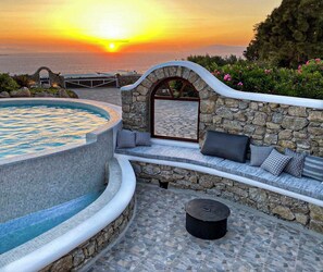 A pool with a stone wall and seating area, a stone bench with cushions, and a view of the sunset over the sea. - Stunning Mykonos Villa Delos Sunset View -close to beaches and town! (Mykonos)