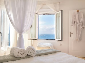Ein Schlafzimmer mit Bett, einem Fenster mit Meerblick, einem an der Wand hängenden Bademantel und einem auf dem Bett liegenden zusammenge rollten Handtuch.