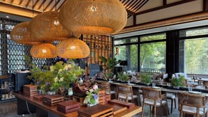 Un restaurant avec des tables et des chaises en bois, des lampes en osier suspendues au plafond et une vue sur les arbres à travers de grandes fenêtres.