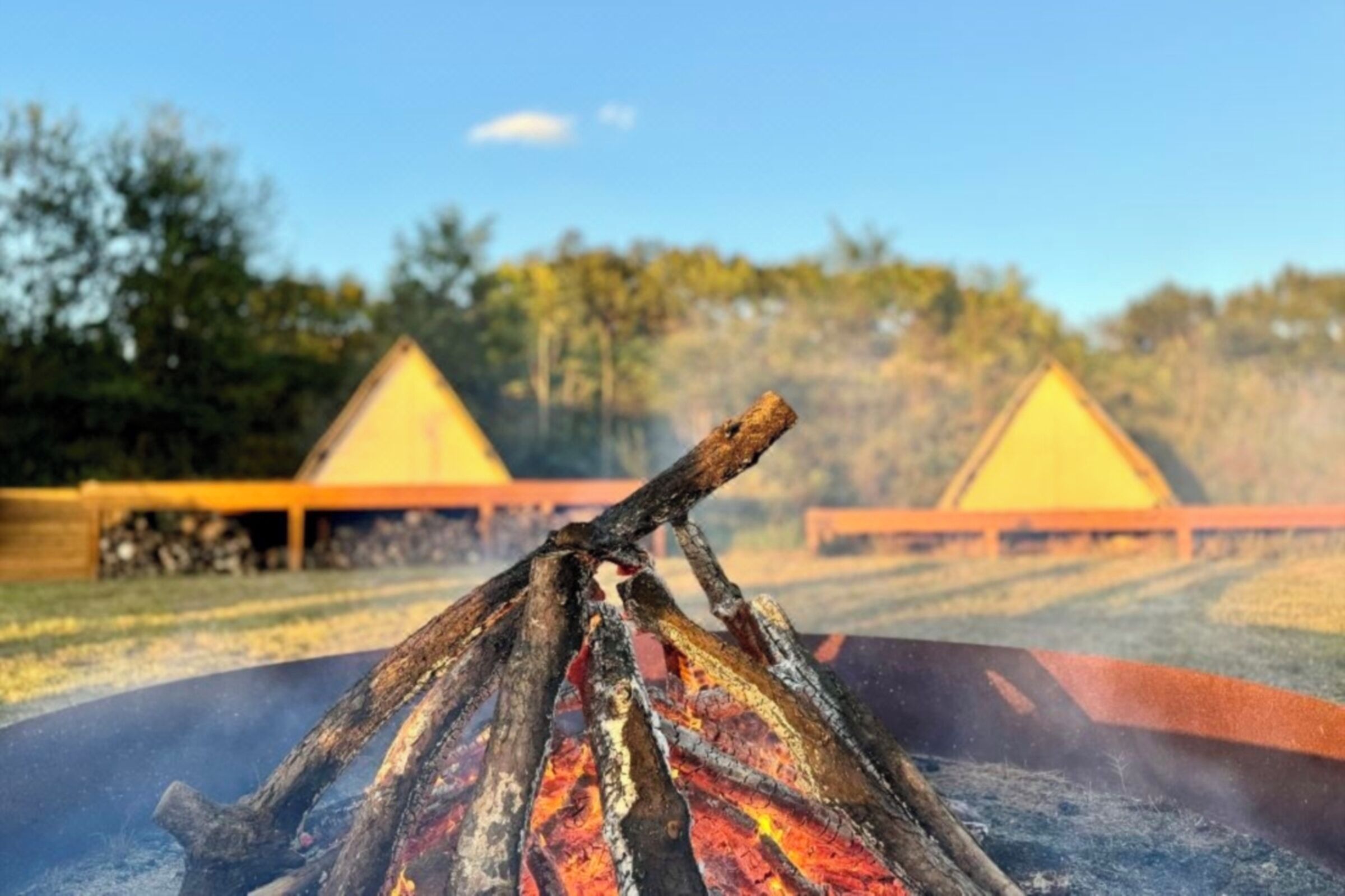 A campfire with logs and embers, surrounded by a circular fire pit. In the background, there are two yellow-roofed structures, possibly cabins or tents, set against a backdrop of trees and a clear sky.
