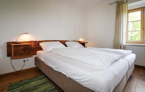 Een tweepersoonsbed met witte beddengoed, een houten hoofdbord, nachtlampjes, een raam met gordijnen en een radiator.