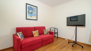 Ein rotes Sofa mit bunten Kissen, ein gerahmtes Bild an der Wand und ein Fernseher auf einem Schrank.