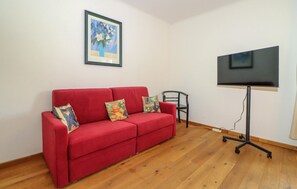 Ein rotes Sofa mit bunten Kissen, ein gerahmtes Bild an der Wand und ein Fernseher auf einem Schrank.