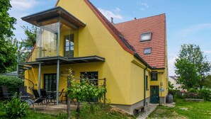 Ein gelbes, zweistöckiges Haus mit rotem Dach, einem Balkon und einem kleinen Garten mit Sitzgelegenheiten.