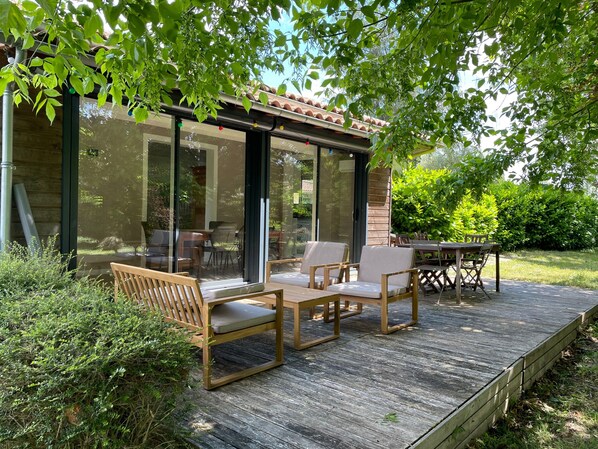 Une terrasse en bois avec des meubles dâextĂ©rieur, un patio fermĂ© par des baies vitrĂ©es et une vue sur la verdure environnante.