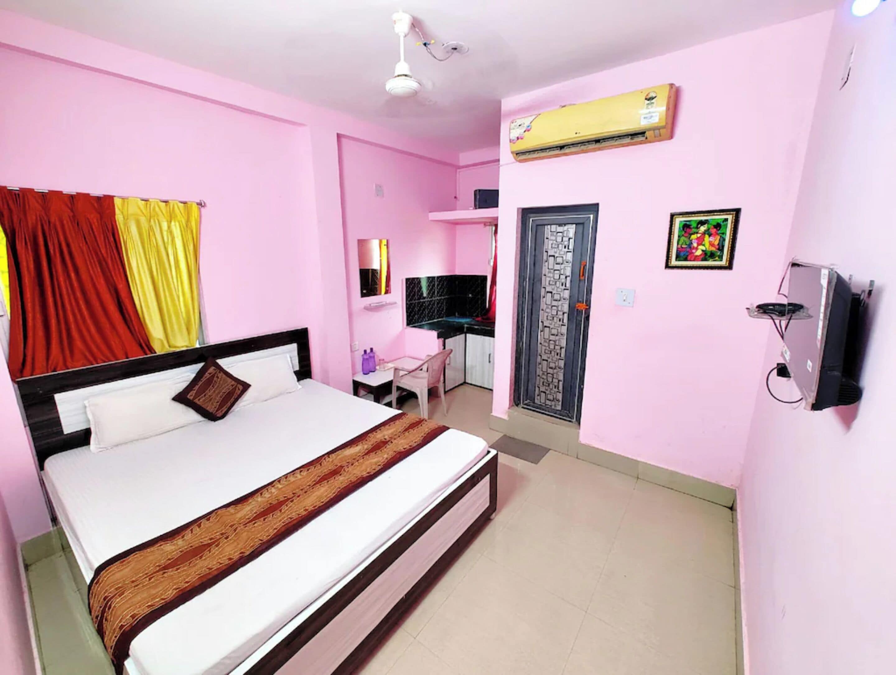 A pink-themed hotel room with a bed, a desk, an air conditioning unit, and a door with a decorative pattern.