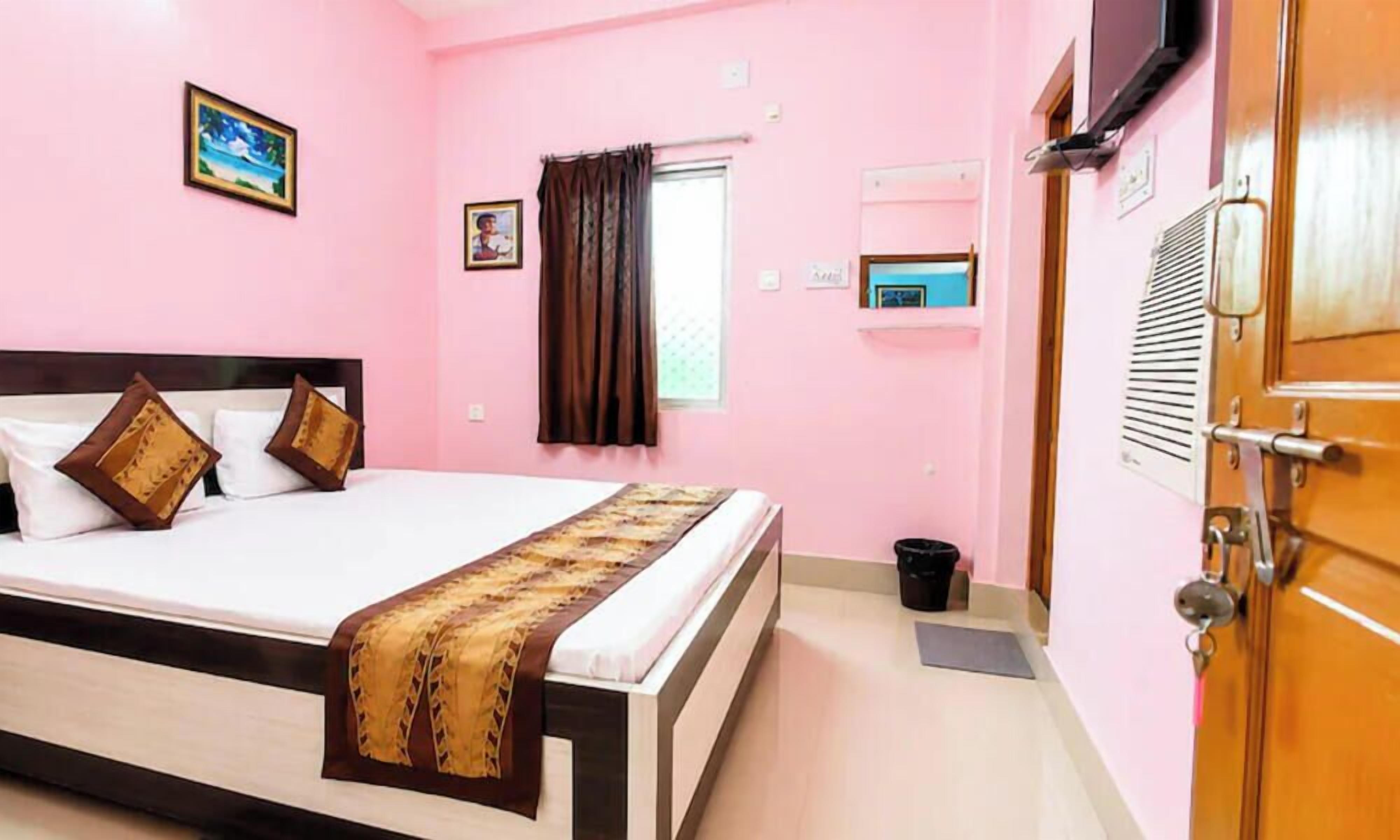A pink-walled room with a bed, a television, a window with curtains, and a door.