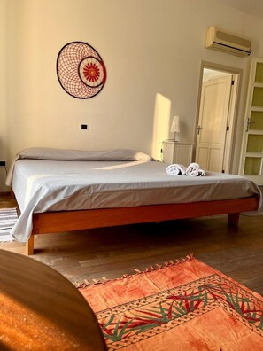 A single bed with a white mattress and a wooden frame, a wall-mounted air conditioner, and a patterned rug on the floor.