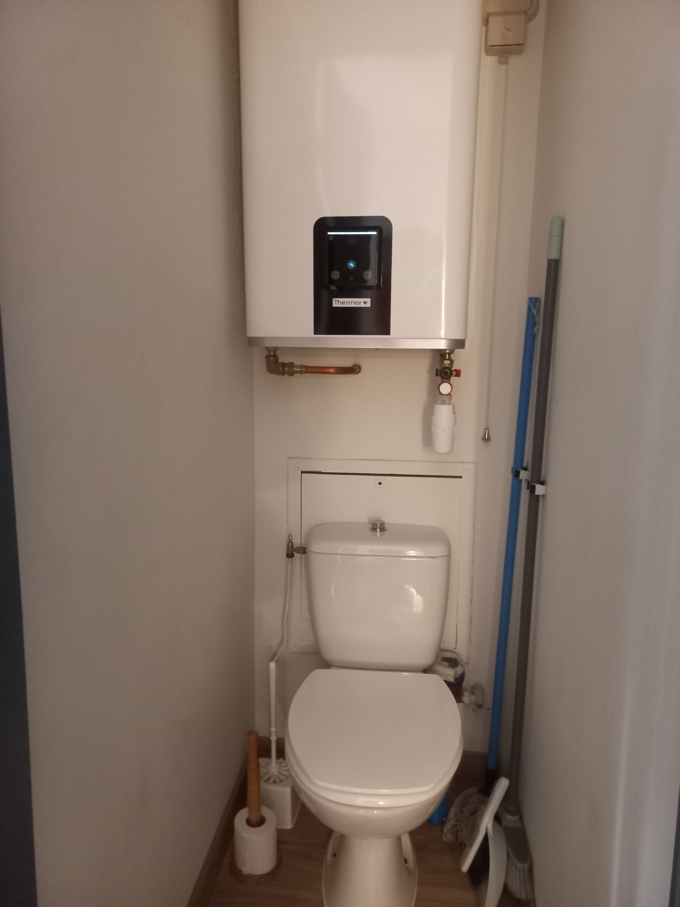 Une salle de bain avec des toilettes blanches, un chauffe-eau apparent et un balai appuyé contre le mur.