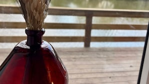 Un florero rojo de vidrio con hierba seca sobre una terraza de madera con vista a un cuerpo de agua.