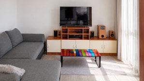 A living room with a grey sofa, a wooden entertainment unit with a TV, speakers, and a colorful coffee table. - Nice apartment in Jesenice with lake view (Jesenice)