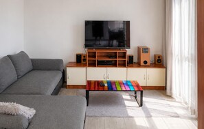 A living room with a grey sofa, a wooden entertainment unit with a TV, speakers, and a colorful coffee table. - Nice apartment in Jesenice with lake view (Jesenice)