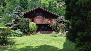 A wooden cabin with a balcony, surrounded by lush greenery and a forested hillside. - Chalet Cosy et Nature avec Grand Jardin & Vue Montagne | Ideal for Family and Friends (Le Ménil)
