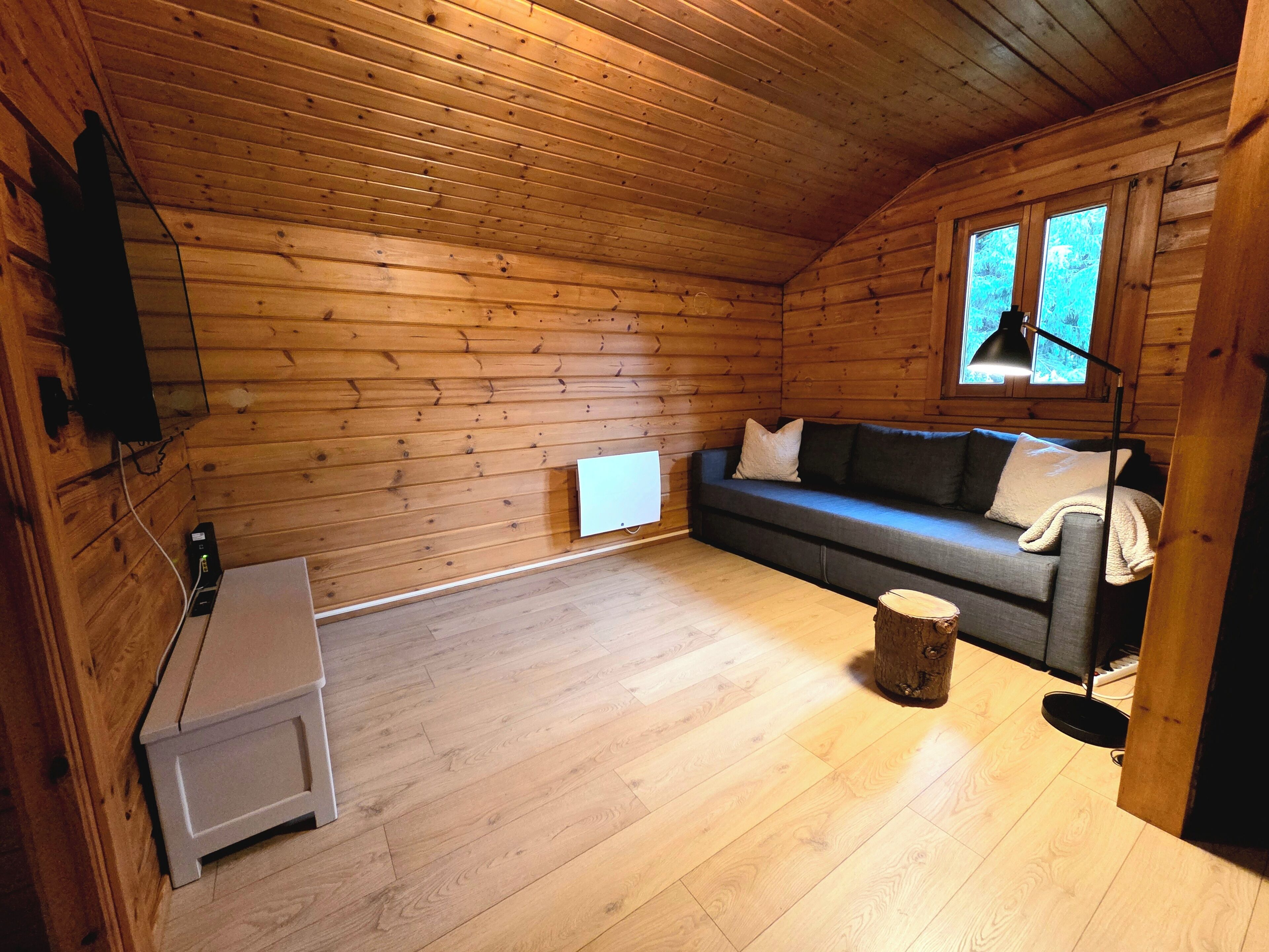 L’intérieur d’une cabane en rondins, avec un plancher en bois, un canapé gris agrémenté de coussins, une petite table et une fenêtre avec une lampe.