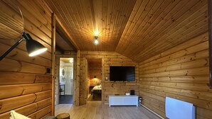 A log cabin interior with a wooden ceiling, a TV mounted on the wall, a white dresser, and a sofa with cushions. - Chalet Cosy et Nature avec Grand Jardin & Vue Montagne | Ideal for Family and Friends (Le Ménil)