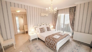 Ein Schlafzimmer mit einem großen Bett, einer Kristallleuchter, einem Fenster mit Vorhängen, einer Heizung und einem kleinen Tisch mit einer Lampe.
