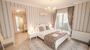 Ein Schlafzimmer mit einem großen Bett, einer Kristallleuchter, einem Fenster mit Vorhängen, einer Heizung und einem kleinen Tisch mit einer Lampe.