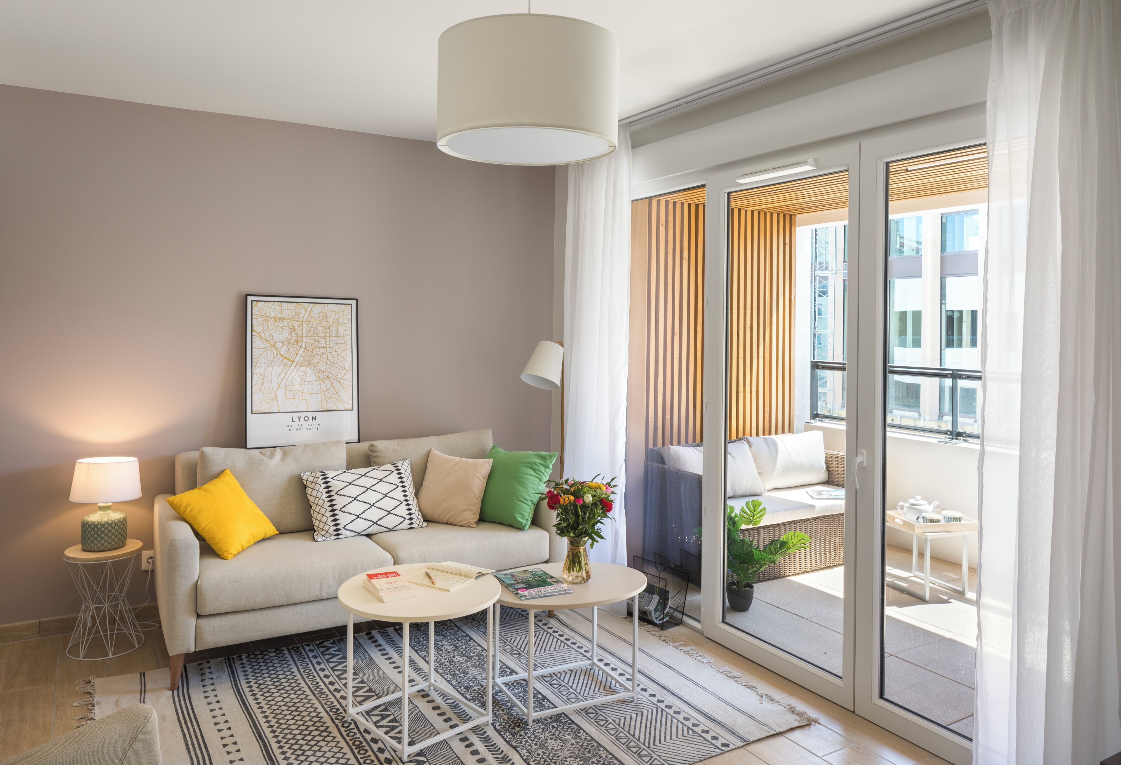A modern living room with a sofa, a small round table, a lamp, and a large window with blinds.