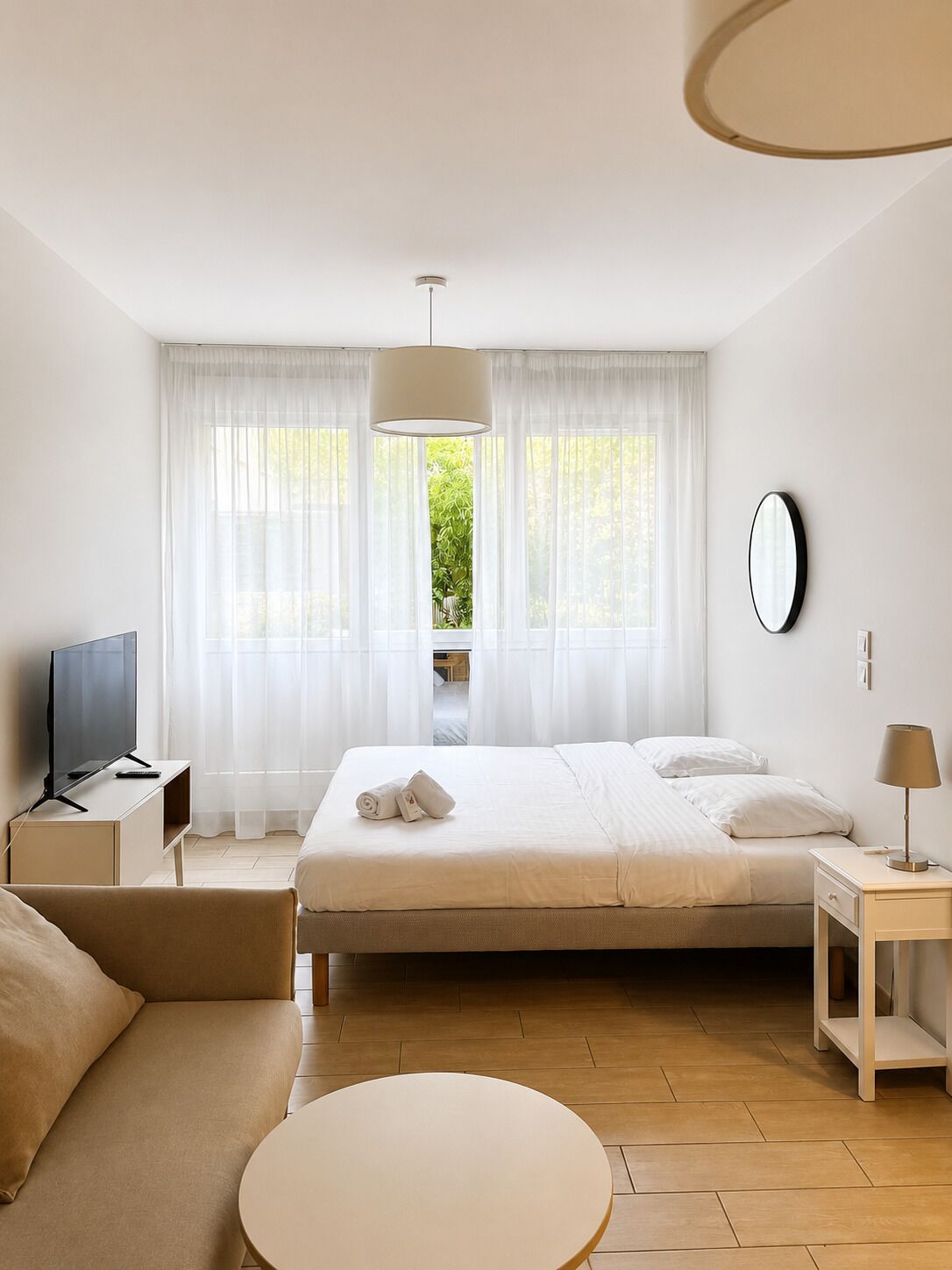 A hotel room with a bed, a TV, a sofa, a round table, and a view of greenery outside.
