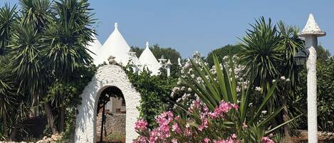 Un bâtiment blanc orné d’une arche en pierre, entouré d’une végétation luxuriante et de fleurs roses.