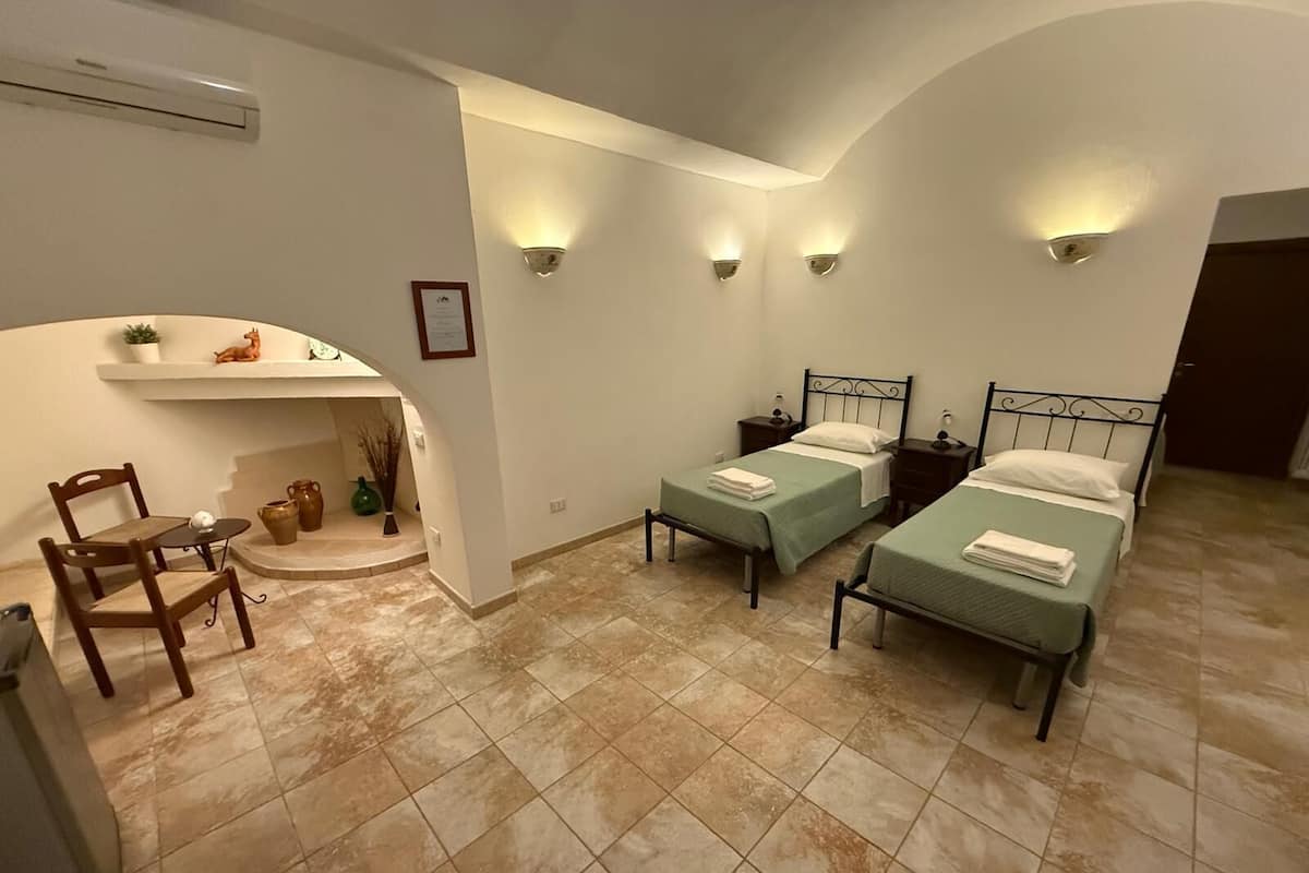 A room with two beds, a small table with chairs, a shelf with decorative items, and wall-mounted lights.