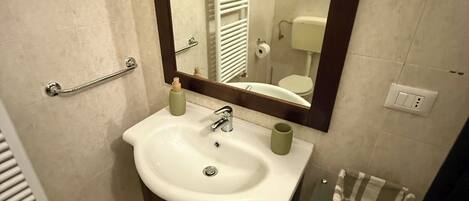 Une salle de bain avec un lavabo blanc, un miroir, un porte-serviettes et une étagère en verre sur laquelle se trouve un panier.