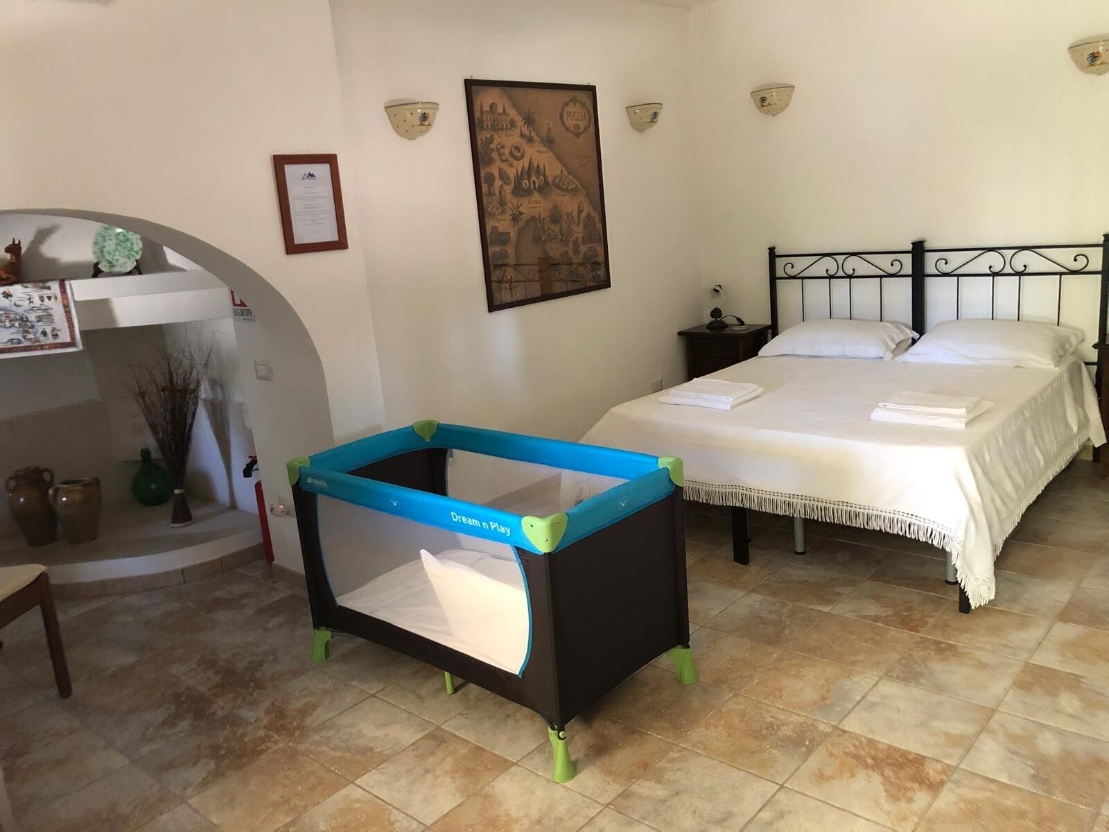 A bedroom with a bed, a crib, a nightstand, a wall-mounted lamp, and a framed picture.
