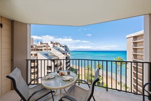 A balcony with a table and chairs, overlooking a beach and ocean. - Ocean-view 1BR condo with pool, lanai, and full kitchen- Valley Isle 1001 (Lahaina)