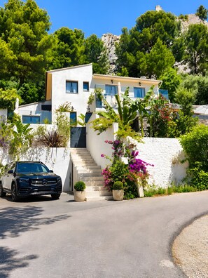 A modern two-story house with a car parked in front, surrounded by lush greenery and flowering plants.
