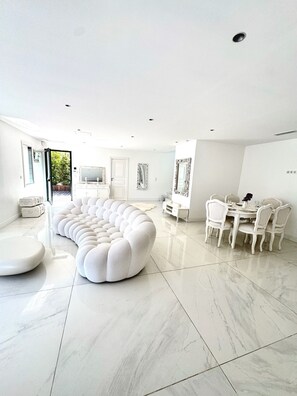 A spacious living area with a large white sectional sofa, a dining table with chairs, and a kitchen area with white cabinetry.
