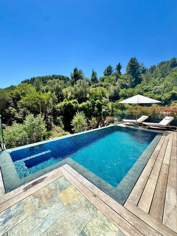 A rectangular swimming pool with a wooden deck and lounge chairs, surrounded by lush greenery and trees.